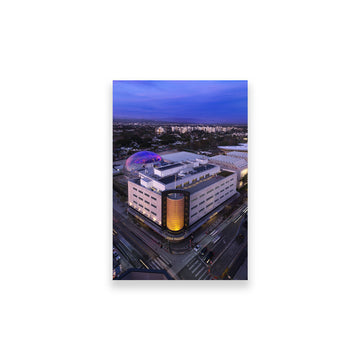 Modern building with a unique design at dusk, surrounded by a cityscape. Academy Museum Postcard Aerial View The Gold Cylinder Facade Size 4 x 6in. Academy Museum Store Exclusive  
©Academy Museum Foundation, Photo by: Fredrik Nilsen